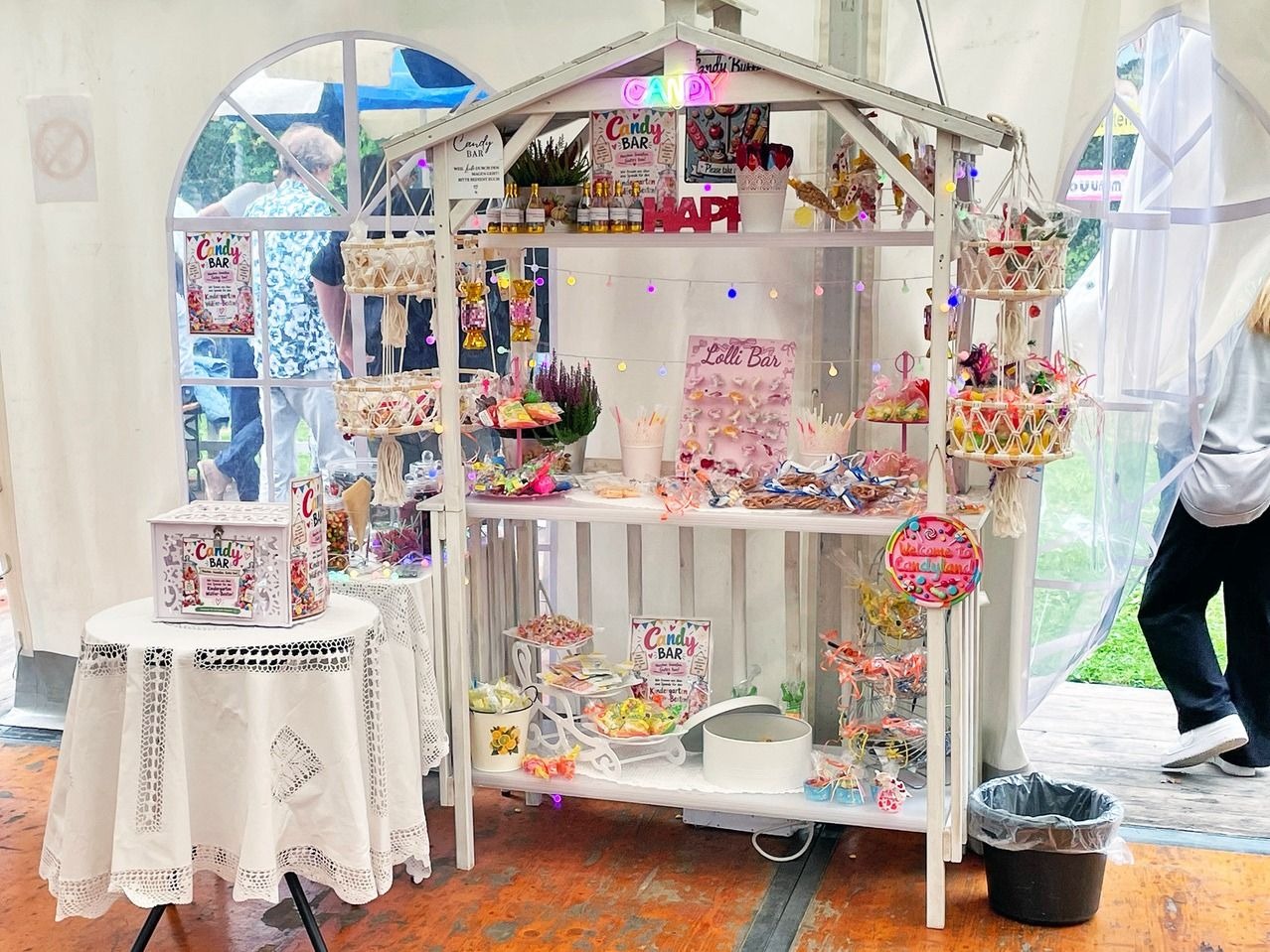 Weißer Süßigkeitenstand mit Bonbons, Lollis, kleinen Flaschen und Dosen sowie bunter Lichterkette in einem Festzelt