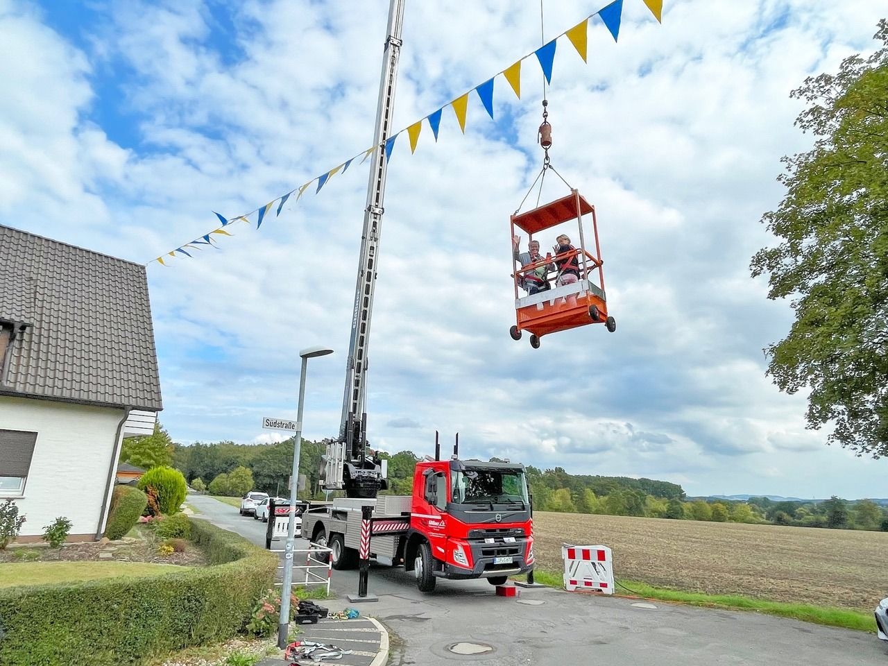 Roter Kranwagen hebt zwei winkende Personen in einem Korb über blau-gelben Festgirlanden an der Südstraße