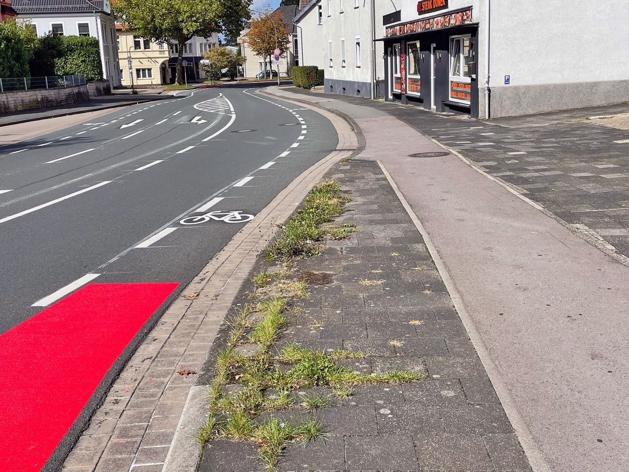 Kurvige Straße mit weißen Radfahrstreifen, einem roten Radwegabschnitt und Bürgersteig vor einem Dönerladen