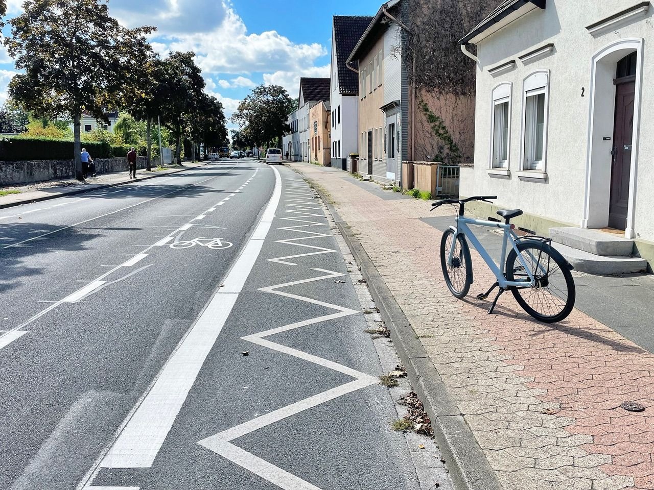 Helles Fahrrad auf Bürgersteig vor weißem Haus, daneben Straße mit Radfahrstreifen und Zickzack-Markierungen