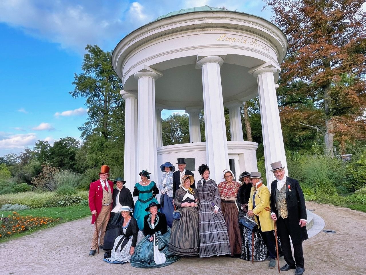 Mehrere Personen in viktorianischer Kleidung posieren vor dem weißen Leopold-Sprudel Pavillon im Park