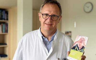 Mann in weißem Kittel und Brille hält Broschüre zur 10. Lippischen Parkinson-Akademie