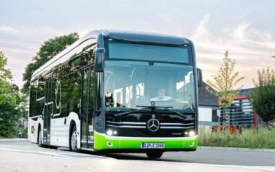 Schwarz-weiß-grüner Stadtbus mit einem Fahrer am Steuer auf einer Straße vor Bäumen und Gebäuden