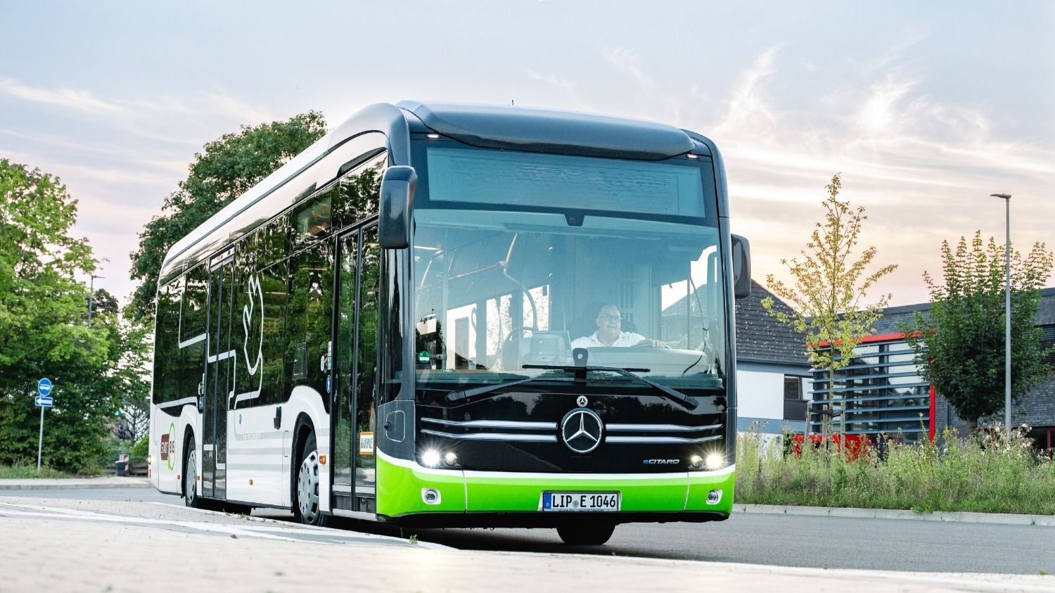 Schwarz-weiß-grüner Stadtbus mit einem Fahrer am Steuer auf einer Straße vor Bäumen und Gebäuden
