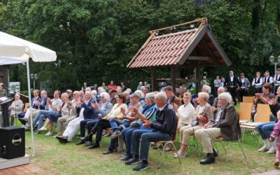 Redner spricht auf einer Bühne vor applaudierendem Publikum, Musikern und Trachtengruppe bei einem Fest im Grünen