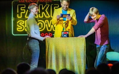 Drei Personen auf der Bühne einer Quizshow: Ein Moderator und zwei Kandidaten vor einem Leuchtschriftzug "Fake Show