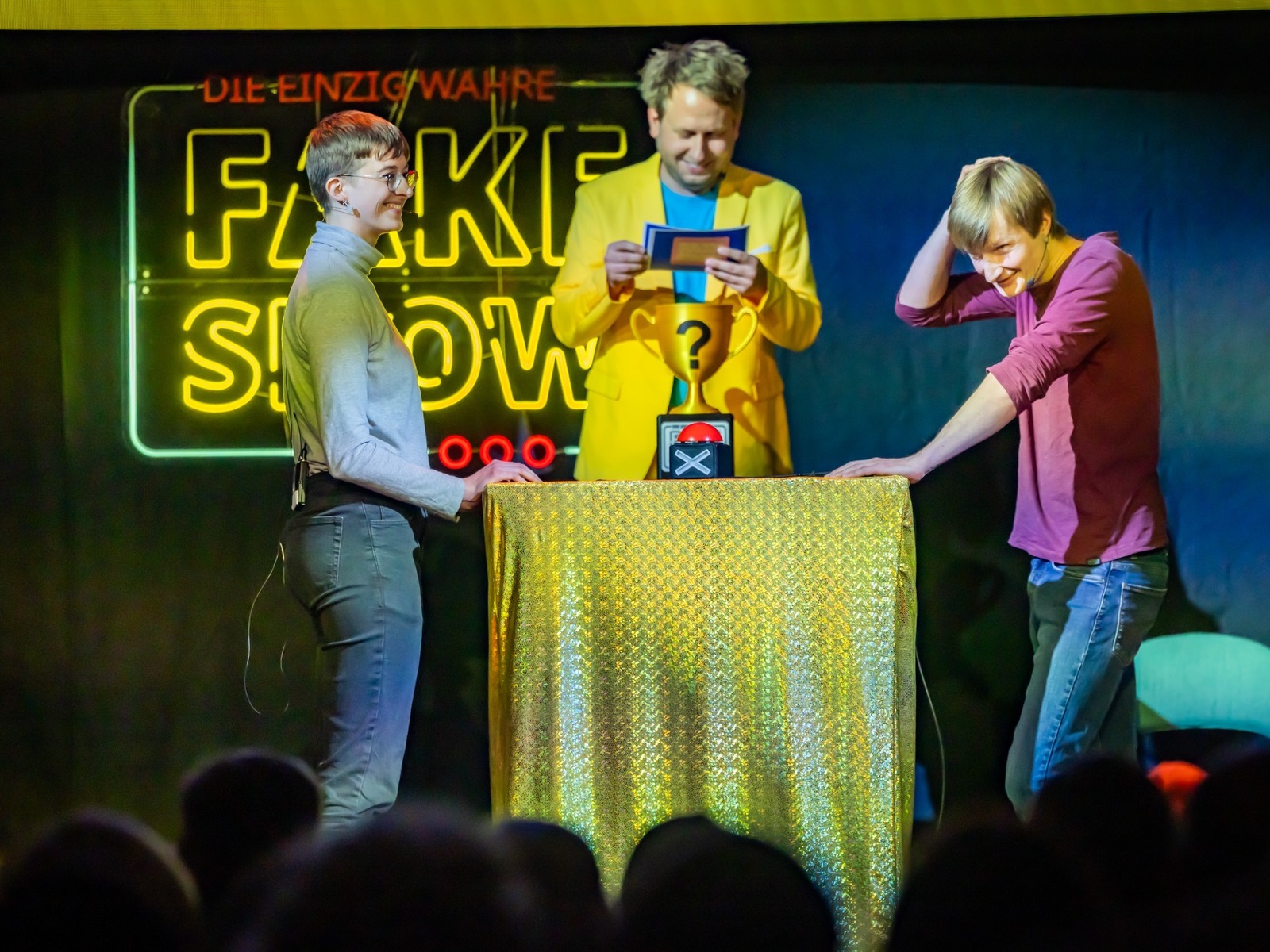 Drei Personen auf der Bühne einer Quizshow: Ein Moderator und zwei Kandidaten vor einem Leuchtschriftzug 