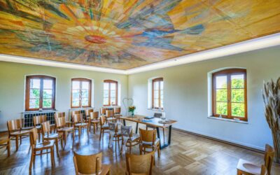 Großer Saal mit farbigem Deckengemälde, Holzstühlen in Reihen mit Mr/Mrs-Schildern, Fenstern und dekoriertem Tisch