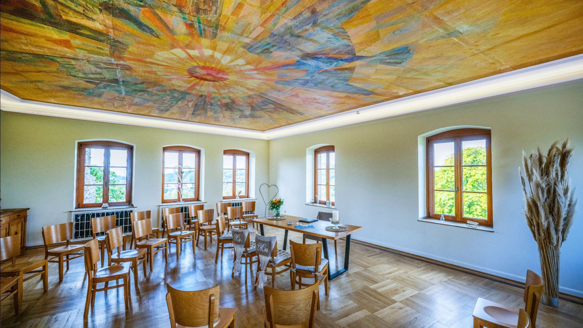 Großer Saal mit farbigem Deckengemälde, Holzstühlen in Reihen mit Mr/Mrs-Schildern, Fenstern und dekoriertem Tisch