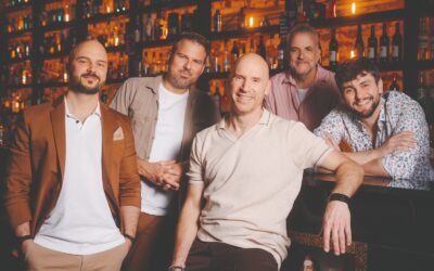 Fünf lächelnde Männer von Alte Bekannte posieren in einer Bar vor einem beleuchteten Regal mit Flaschen