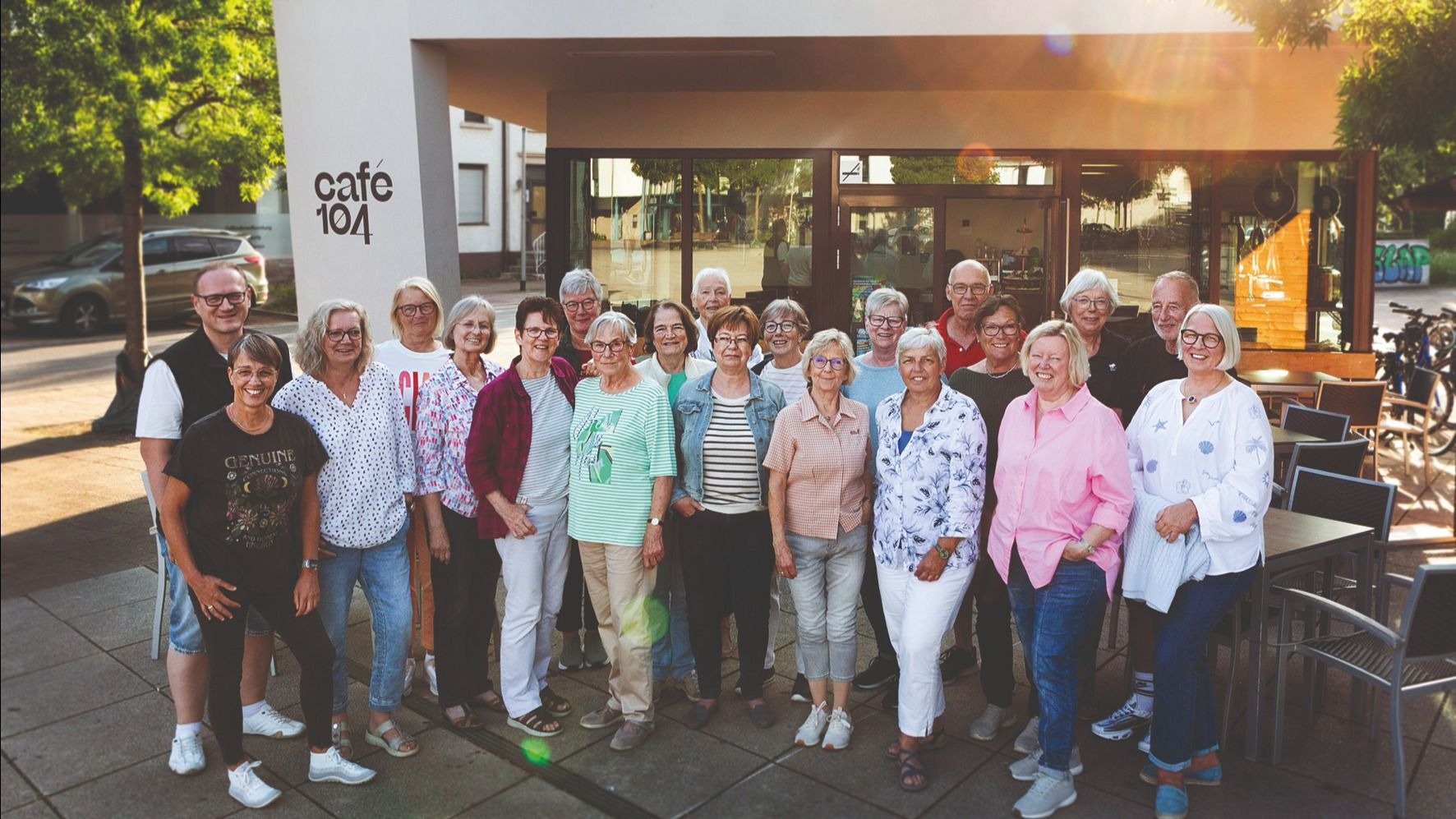 20 lächelnde Erwachsene posieren als Gruppe vor dem Café 104 mit seinen Außentischen
