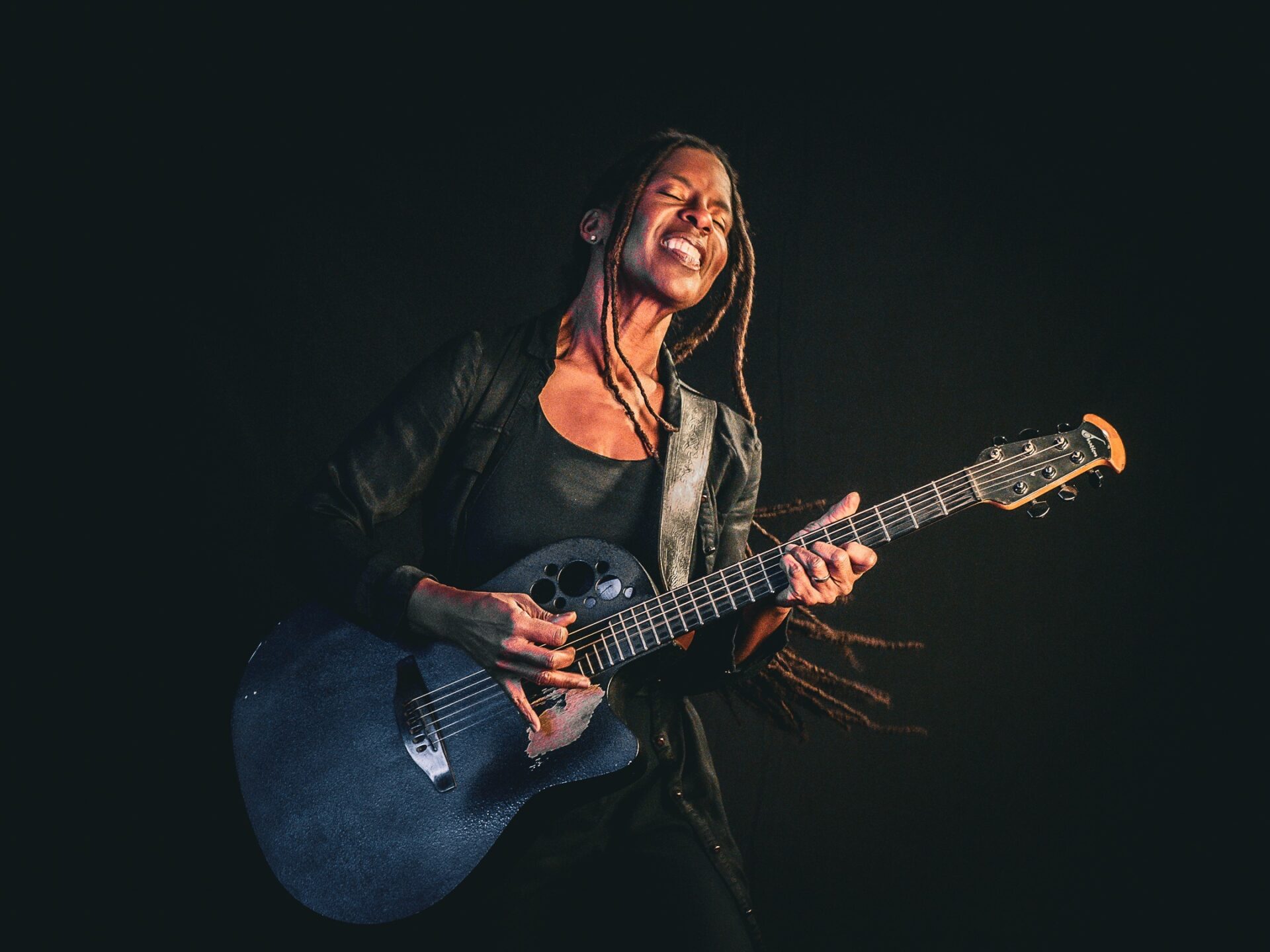 Judy Bailey spielt mit geschlossenen Augen und geöffnetem Mund eine dunkelblaue Akustikgitarre auf dunklem Hintergrund