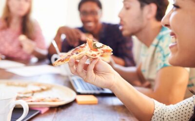 Eine lachende Frau hält ein Pizzastück vor dem Mund, im Hintergrund essen weitere Menschen an einem Tisch