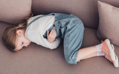 Kleines Mädchen mit Zopf in blauer Latzhose liegt gekrümmt auf der Seite auf einem beigen Sofa