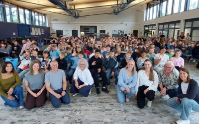 Viele winkende Fünftklässler und lächelnde Erwachsene sitzen in einer großen Schulaula für ein Gruppenfoto