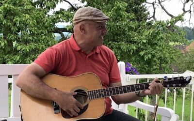 Ennio spielt lächelnd Akustikgitarre im Freien vor grünen Bäumen auf einer weißen Bank