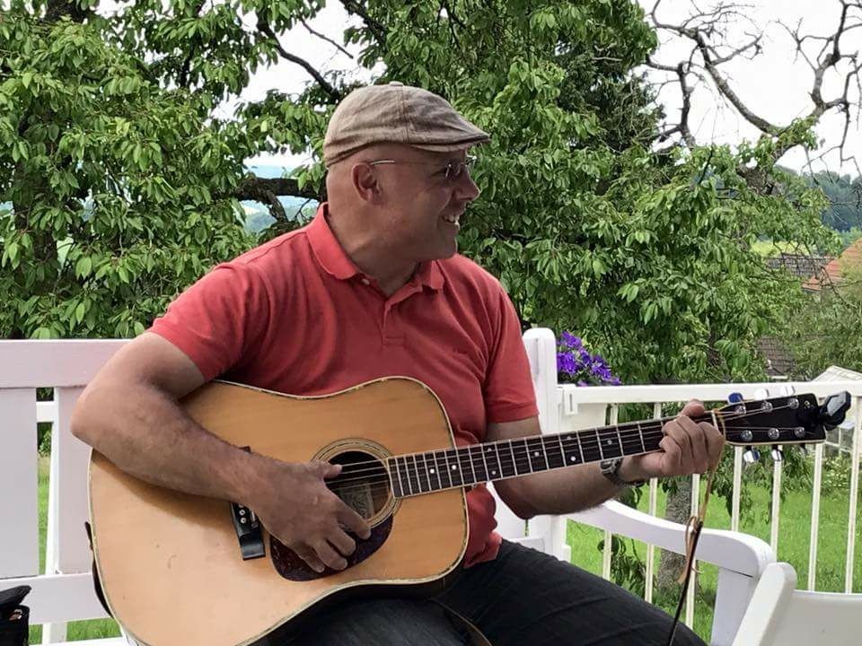 Ennio spielt lächelnd Akustikgitarre im Freien vor grünen Bäumen auf einer weißen Bank