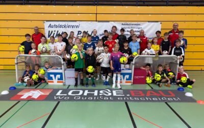 Lächelnde Kinder und Erwachsene in Fußballtrikots posieren mit Bällen in einer Sporthalle vor Bannern