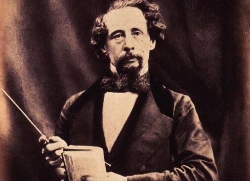 Charles Dickens mit Bart und Schnurrbart, einen Notizblock haltend und mit einem Zeigestab darauf zeigend