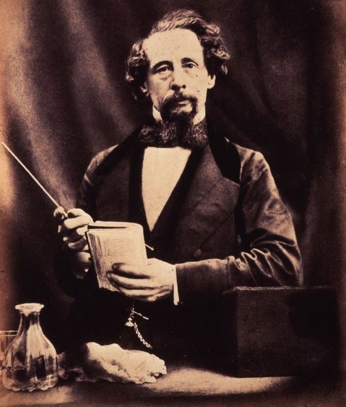 Charles Dickens mit Bart und Schnurrbart, einen Notizblock haltend und mit einem Zeigestab darauf zeigend