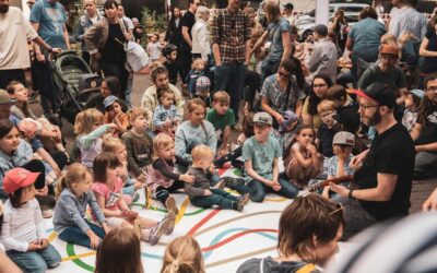 Kinder und Erwachsene sitzen auf einer weißen Matte mit bunten Linien und sehen einem Gitarrenspieler zu