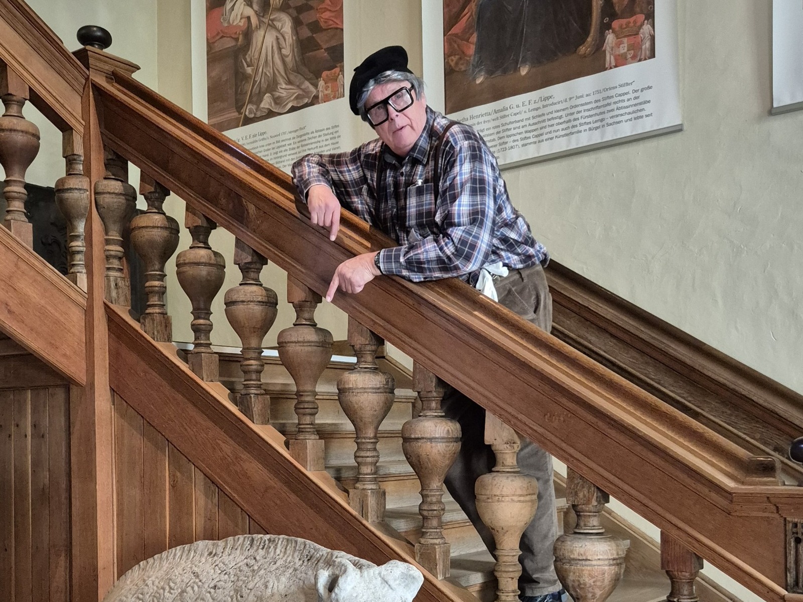 Gustav Grummel mit Beret und Brille lehnt an einer Holztreppe, zeigt nach unten, vor historischen Porträts und einer