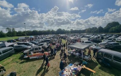 Großer Flohmarkt auf einer Wiese mit vielen Autos, Marktständen und Menschen unter blauem Himmel mit Wolken in
