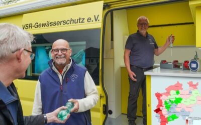 Zwei Männer übergeben und empfangen eine Wasserflasche am gelben VSR-Gewässerschutz Labormobil, ein Dritter steht mit
