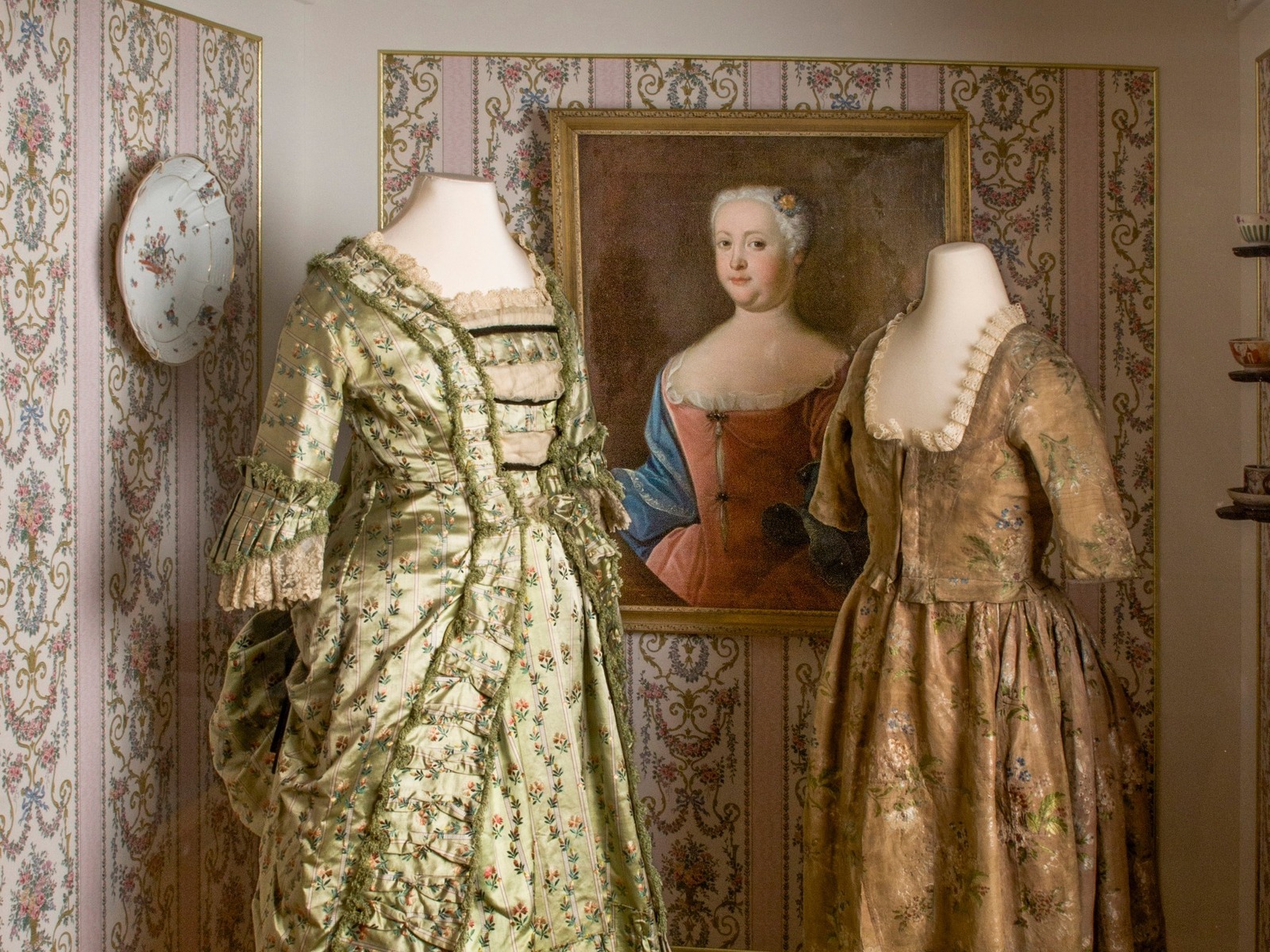 Zwei historische Kleider auf Schneiderpuppen, ein Frauenporträt, ein Fächer, Schuhe und ein Dekoteller vor gemusterter