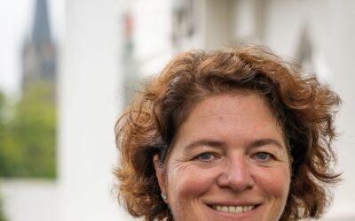 Kerstin Vieregge lächelt mit lockigen, braunen Haaren in dunkelblauer Jacke vor einer Kirche und weißem Gebäude