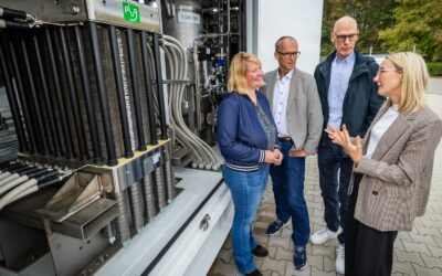 Eine Frau gestikuliert im Gespräch mit drei weiteren Personen vor einem Elektrolyseur mit Rohren und dem "Plug"-Logo