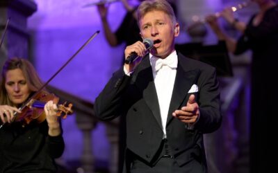 Helmut Lotti singt in schwarzem Smoking mit Mikrofon auf einer Bühne, umgeben von Musikerinnen mit Violine und Querflöte