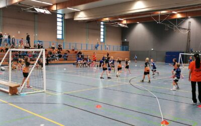 Kinder spielen Handball in einer Sporthalle mit Zuschauern auf den Tribünen und einem Erwachsenen am Spielfeldrand