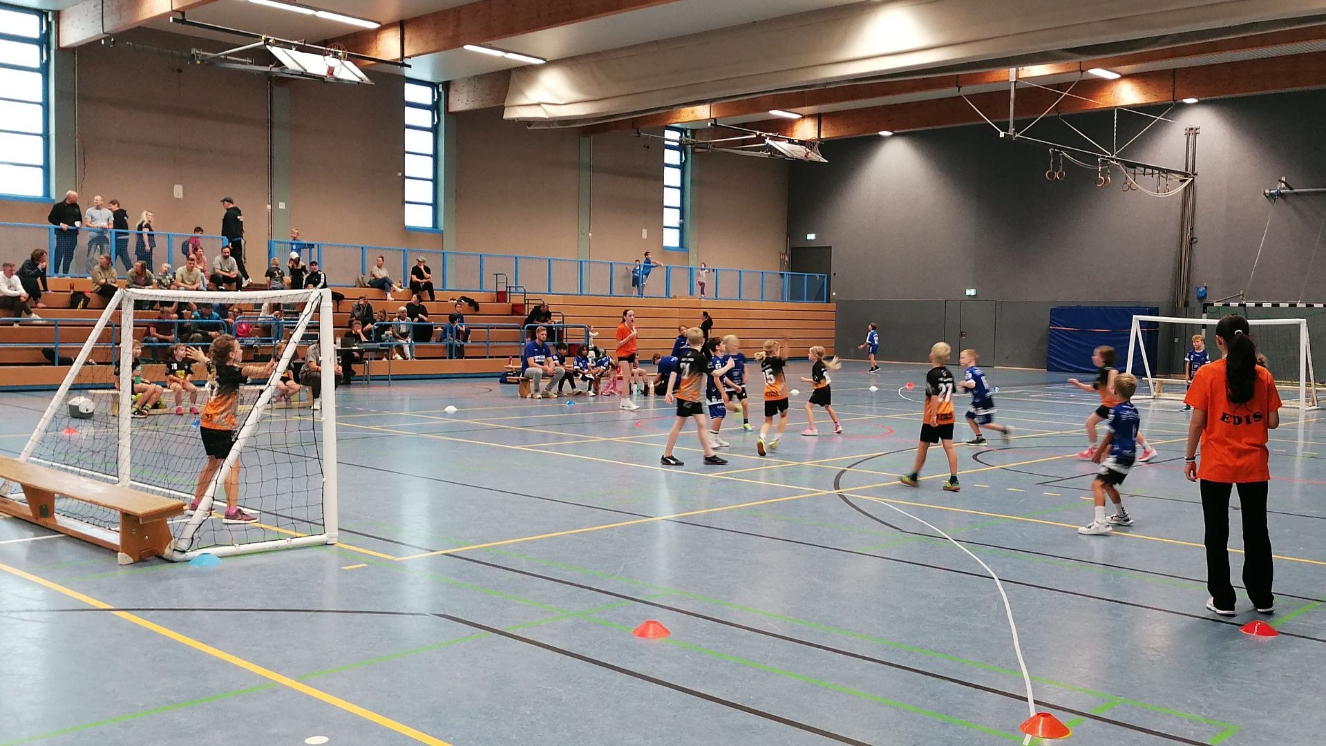Kinder spielen Handball in einer Sporthalle mit Zuschauern auf den Tribünen und einem Erwachsenen am Spielfeldrand