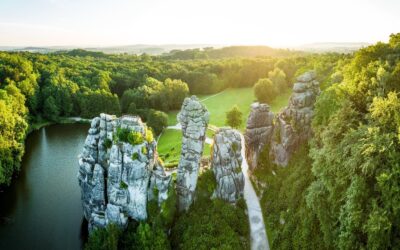 Markante Felsformation der Externsteine am dunklen See, umgeben von dichtem grünen Wald im goldenen Sonnenlicht
