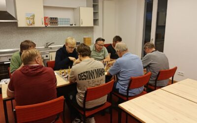Acht Männer spielen konzentriert Schach an zwei Tischen in einem Raum mit Küchenzeile und roten Stühlen