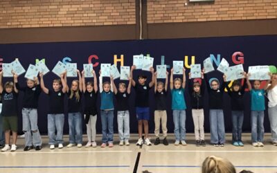 Eine Gruppe von Kindern präsentiert Blätter mit Buchstaben und Illustrationen vor einer blauen Wand mit dem Wort