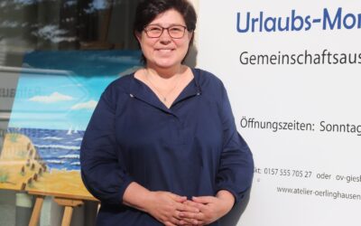 Lächelnde Frau mit Brille vor Schild "Urlaubs-Momente" und Gemälde mit Strand und Meer im Schaufenster
