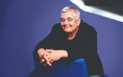 Oschmann lächelt, über die Rückenlehne eines blauen Stuhls gelehnt, vor lilafarbenem Hintergrund