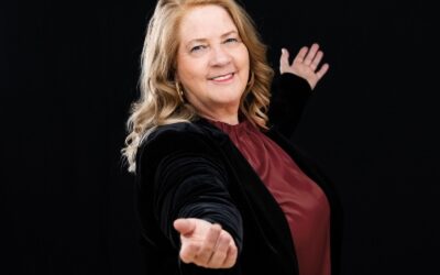 Kathy Kelly lächelt und macht eine einladende Geste mit beiden Armen zum Betrachter