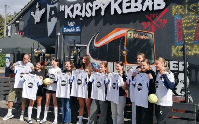 Ein Mann und zehn Mädchen präsentieren neue weiße Trikots mit Sponsorlogo Krispy Kebab vor einem Gebäude mit