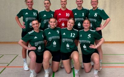 Acht lächelnde Handballspielerinnen in grünen und einem roten Trikot posieren in zwei Reihen in einer Sporthalle