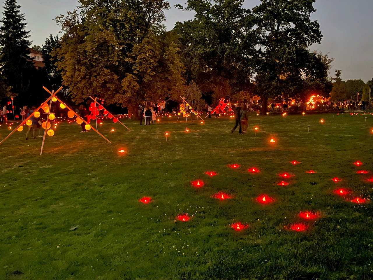 Abendlicher Kurpark, erfüllt von roten Bodenlichtern und bunten Lampions an Holzgestellen, Menschen flanieren zwischen