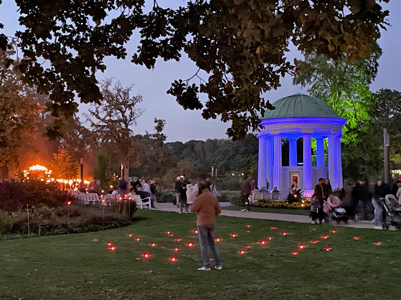 Abendlicher Kurpark mit blau beleuchtetem Pavillon, roten Lichtern auf der Wiese und vielen Besuchern