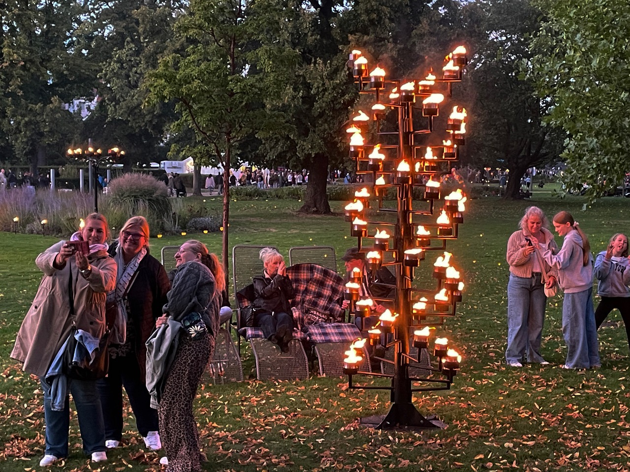 Besucher stehen und sitzen im Dämmerlicht eines Kurparks mit Herbstlaub um eine große, hell leuchtende Flammen-Skulptur