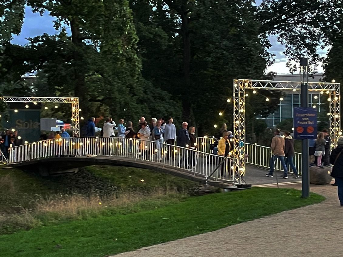 Zahlreiche Besucher auf einer beleuchteten Brücke im Kurpark, flankiert von zwei Lichterbögen bei Dämmerung