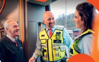 Zwei Sicherheitsleute in gelben Westen mit Bodycams und ein Bahnmitarbeiter lächeln und sprechen in einem Zug