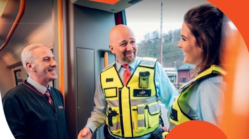 Zwei Sicherheitsleute in gelben Westen mit Bodycams und ein Bahnmitarbeiter lächeln und sprechen in einem Zug