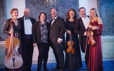 Drei Männer und drei Frauen in festlicher Abendkleidung lächeln mit Cello und zwei Geigen in einem Saal