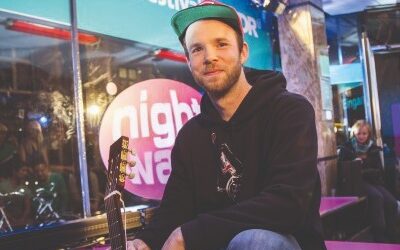 Junger Mann mit Bart, Kappe und dunklem Hoodie sitzt mit einer Akustikgitarre vor WDR-Logo und Menschen